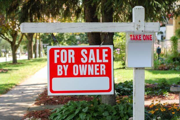 against a suburban backdrop, a red real estate sign with white letters says "for sale by owner"