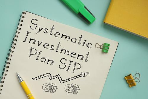 The words &quot;Systematic Investment Plan SIP&quot; appeared drawn in ink on the cover of a spiral notebook surrounded by writing implements an binder clips on a blue tabletop.