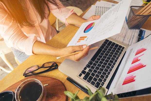 Business woman with long black hair, wearing an orange shirt, holding graph paper and pencil, notebook turn on in front of her and chart performance close a monitor, many document around the table