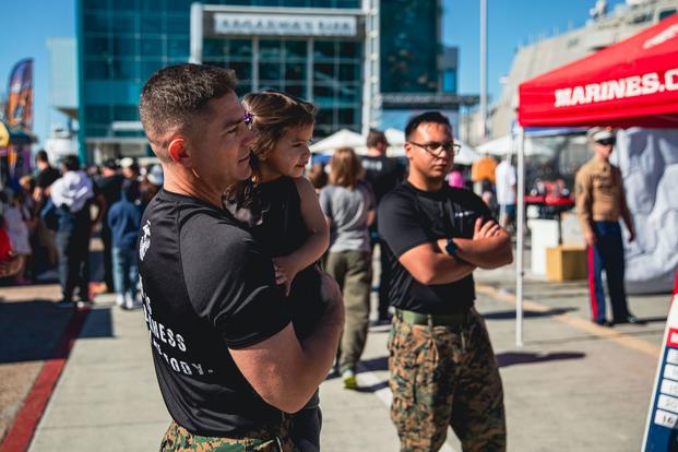 A Marine in cammies and a black T-shirt holds his preschool-age daughter. They both have brown hair.