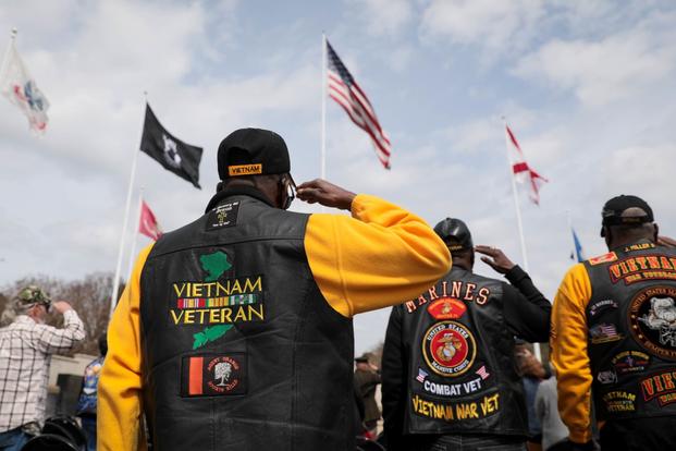 With their backs turned to the camera, Black Vietnam veterans in Vietnam-themed leather jackets decorated with embroidery and patches, salute flags.