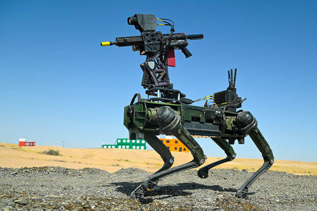 Un "perro robot" cuadrúpedo conocido como Ghost Robotics Vision 60, equipado con una torreta armada en su parte superior. El robot está en un terreno desértico, y la torreta parece tener un rifle de asalto, probablemente un AR-15/M16. Este robot se utiliza en pruebas militares para contrarrestar drones y amenazas terrestres. El fondo muestra un área de entrenamiento con edificios de colores.