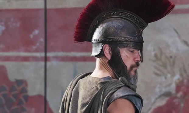 Side profile of Matt Damon wearing a bronze plumed helmet and dark armor as Odysseus in Christopher Nolan’s The Odyssey.