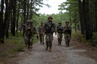 A squad of soldiers in camo walk on a dirt road through the woods toward the camera.