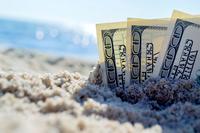 Three US $100 bills stick up from the sand on a beach.