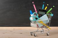 A miniature shopping cart on a wood tabletop contains regular-sized school supplies.